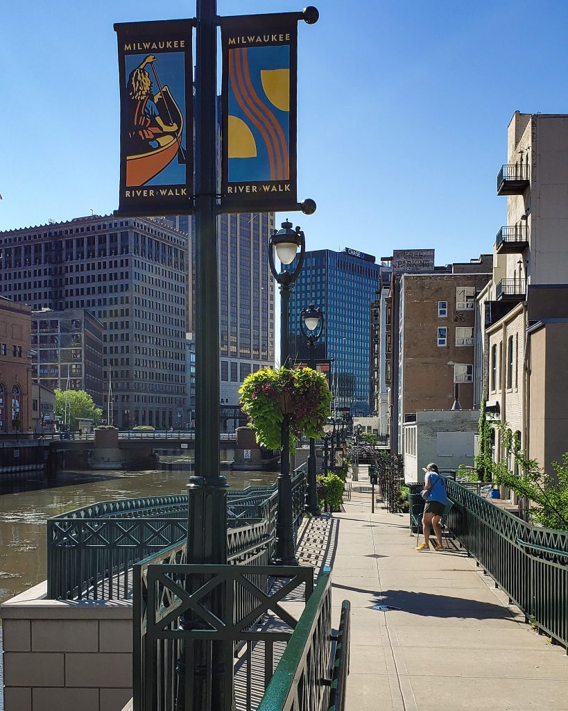 sidewalk next to a river surrounded by city buildings.  View from the Milwaukee River Walk