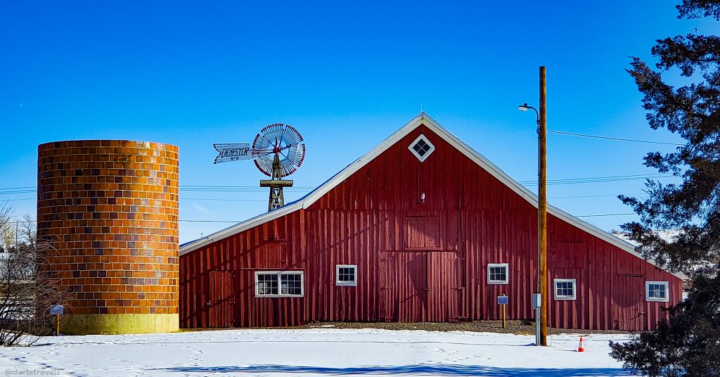 historic red brick silo next to a red barn with sloping roof. Historic windmill in the background.