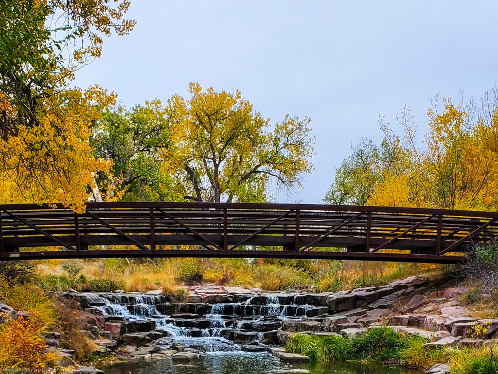 metal pedestrian bridge over a cascading creek with cottonwood trees in the background with yellow and green leaves; near the intersection of the High Line Canal Trail and the Cherry Creek Regional Trail, near Denver