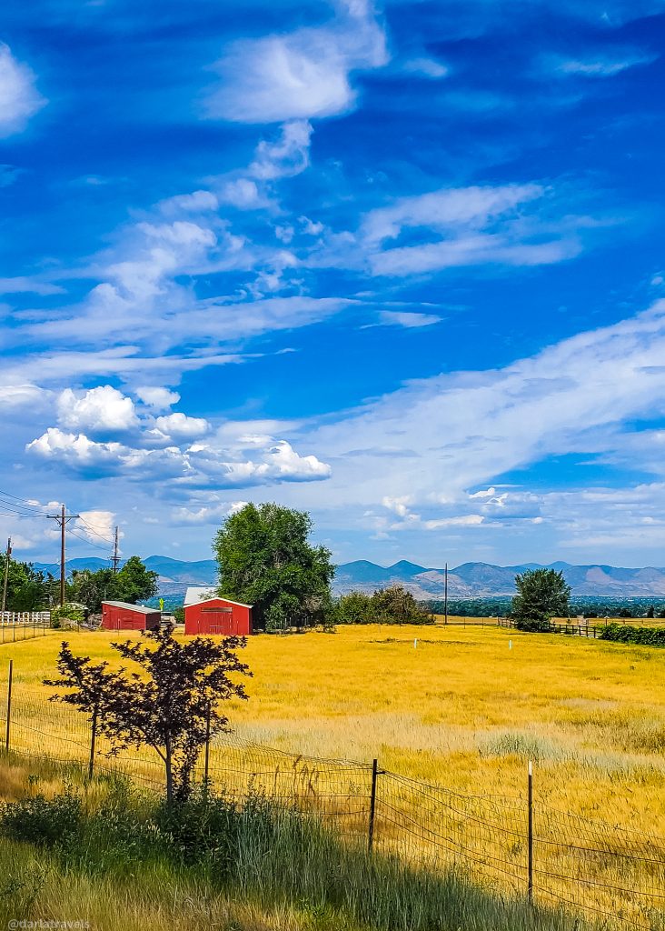 blue sky with clouds, mountain range in the distance.  Midground, red farm buildings with a few green trees, foreground, golden meadow