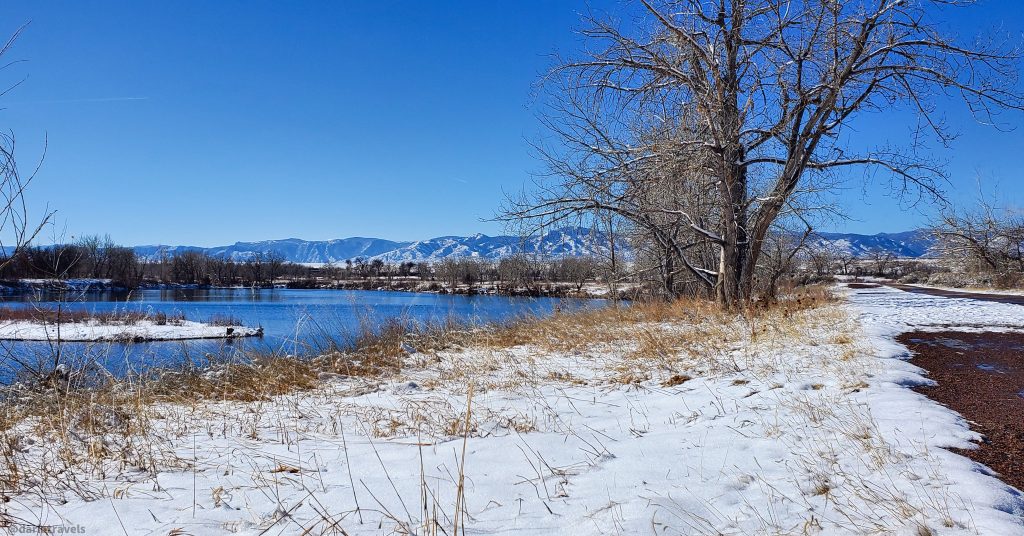 snow covered ground next to paved trail on one side and lake on the other; mountains in the distance.