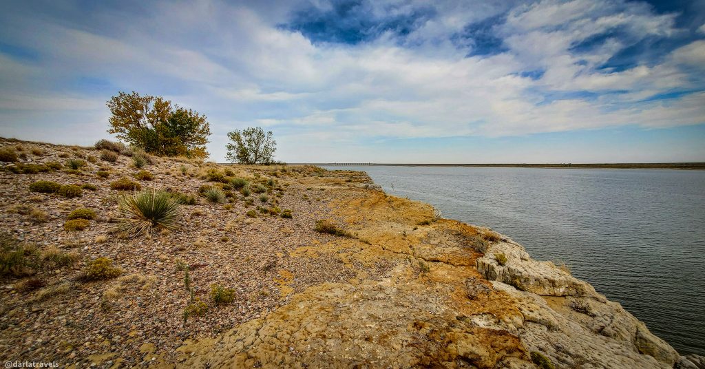 Rocky shoreline of a reservoir with sparse vegetation and a partly cloudy sky. John Martin Dam visible in the distance