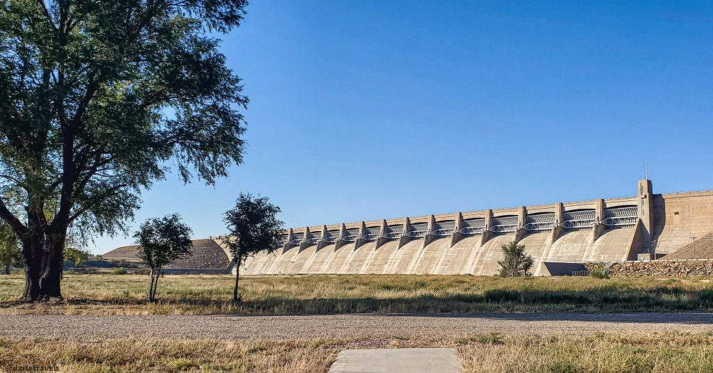 Large concrete dam with numerous sluice gates, situated amongst a grassy landscape and a backdrop of trees on a sunny day.  John Martin Dam, Colorado