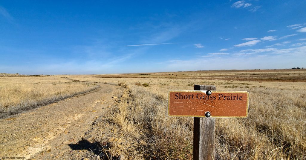 A dirt trail winds through a vast expanse of short grass prairie, marked by a sign reading "Short Grass Prairie". Red Shin Trail, John Martin Reservoir State Park