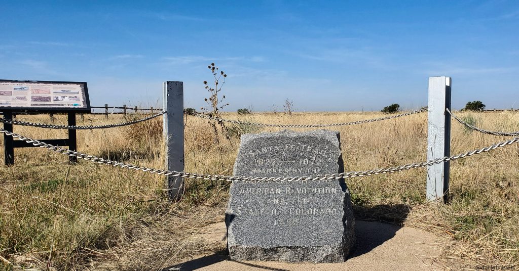 Historical granite marker for the Santa Fe Trail, 1822-1872 marking the American Revolution route through the state of Colorado, surrounded by chain link fence and informational sign. Placed in 1906