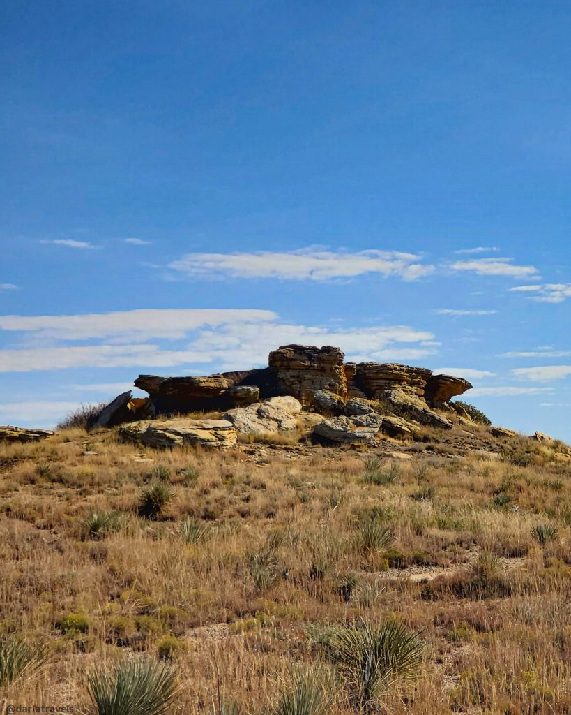 Large, light-tan Dakota sandstone formations rise above a field of dry, light-brown grasses and small desert plants under a clear blue sky.