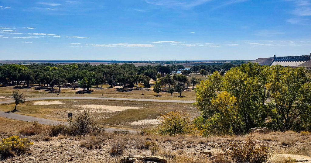 High-angle view of Lake Hasty Campground park-like area with a paved road, trees, and a distant dam.