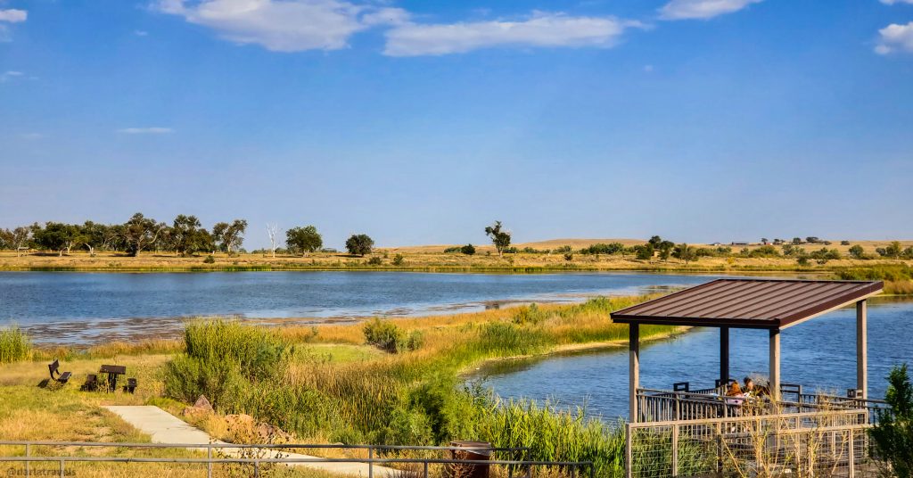 Lakeside gazebo and fishing pier with people enjoying the view, surrounded by grassy wetlands and a calm body of water; Lake Hasty; John Martin Reservoir State Park.