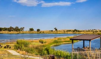 Lakeside gazebo and fishing pier with people enjoying the view, surrounded by grassy wetlands and a calm body of water.