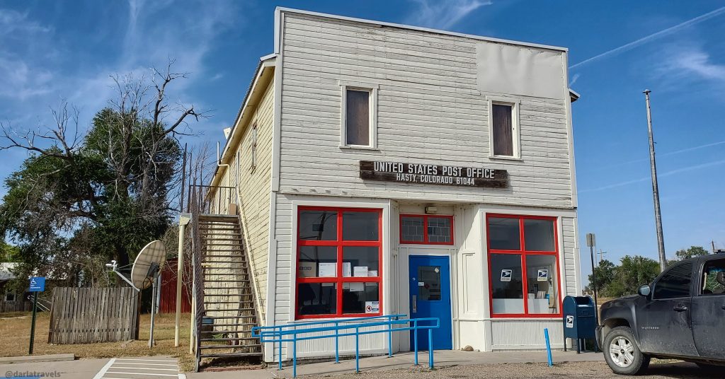 Exterior of a historic, light-beige, two-story post office building in Hasty, Colorado, with red window frames, a blue ramp, and a mailbox.