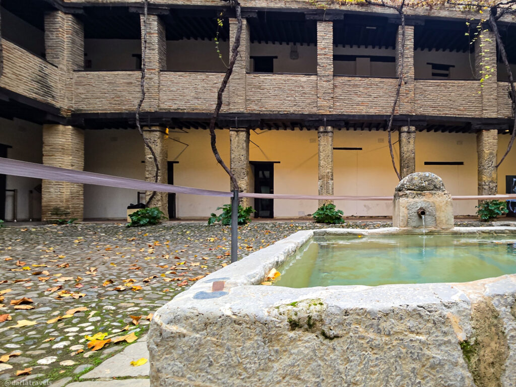 A photo of a courtyard with a small fountain in the center. The courtyard is surrounded by a two-story building with a covered walkway supported by stone columns. The ground is covered with fallen leaves. Corral del Carbon; Part of the Dobla de Oro sites in Granada Spain