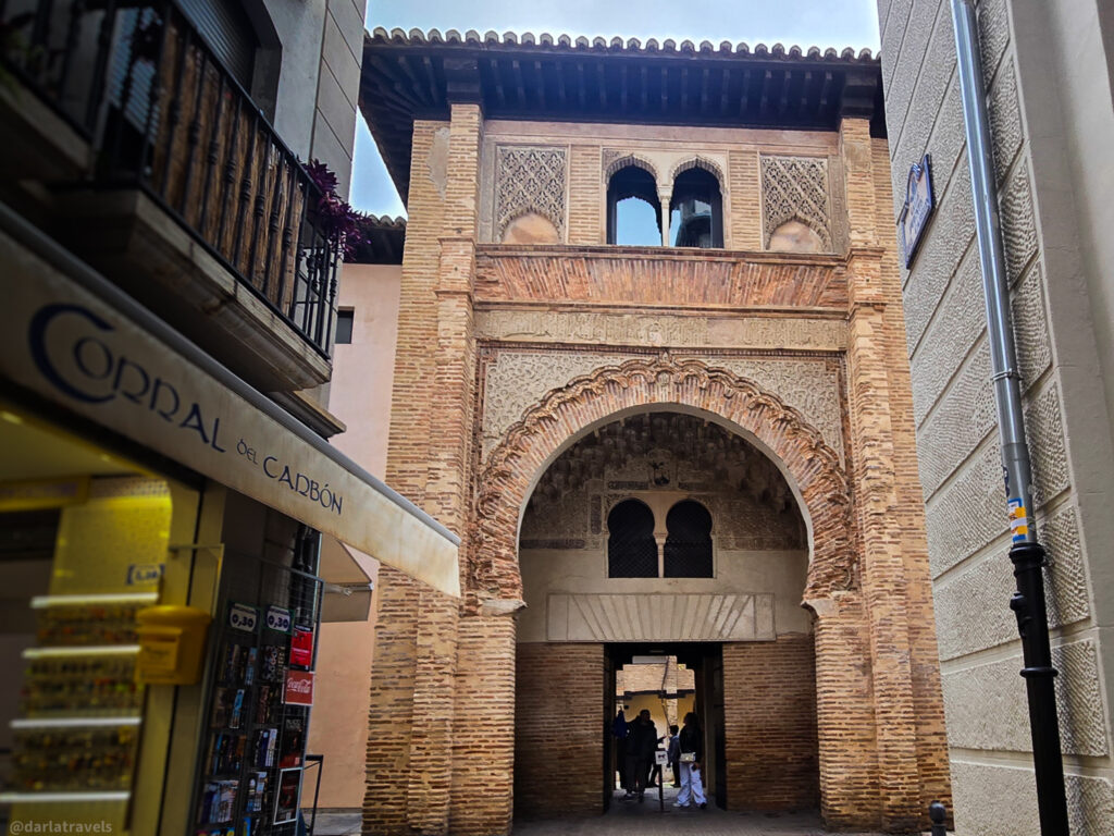 A photo of the entrance to the Corral del CarbĂłn in Granada, Spain. The entrance features a large, elaborately decorated arch with intricate brickwork and Moorish-style details.