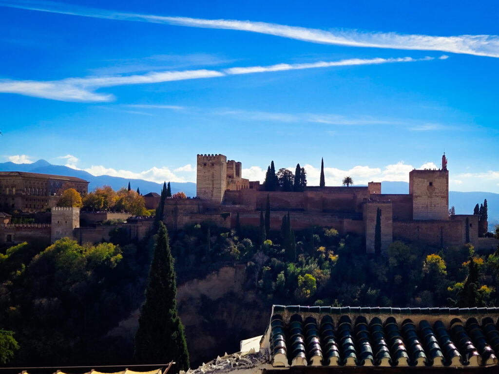 photo showcasing the majestic Alhambra fortress perched atop a hill in Granada. The image captures the fortress's imposing walls, towers, and intricate architecture against a vibrant blue sky with wispy clouds.
