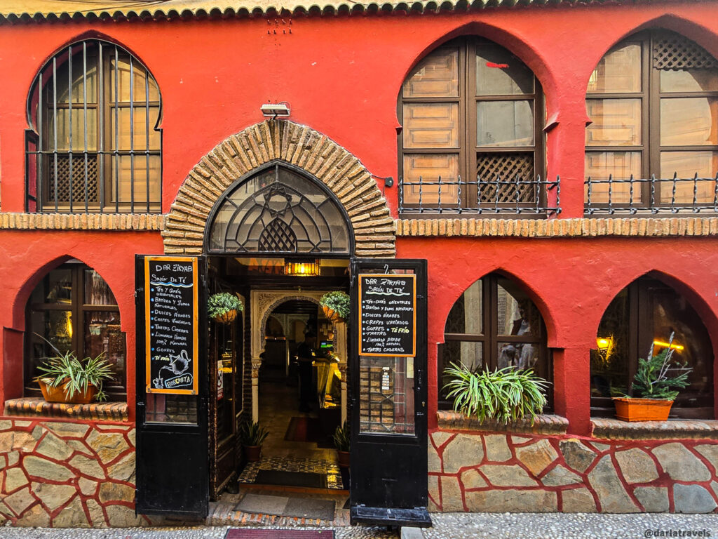 A photo of a building with a red exterior. The building has multiple arched doorways and windows, with potted plants adding a touch of greenery. A tea house.