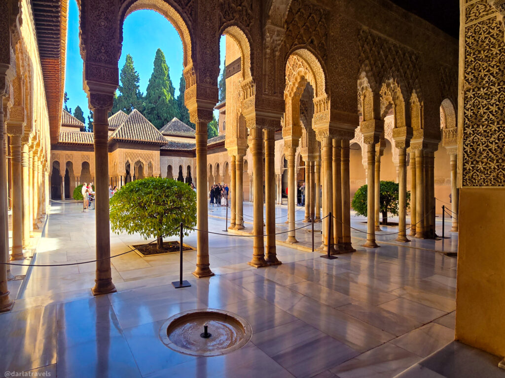 A photo of the Patio de los Leones (Court of the Lions) at the Alhambra in Granada, Spain. The image shows a perspective view through a series of arches with intricate details, leading to a central courtyard with a fountain.