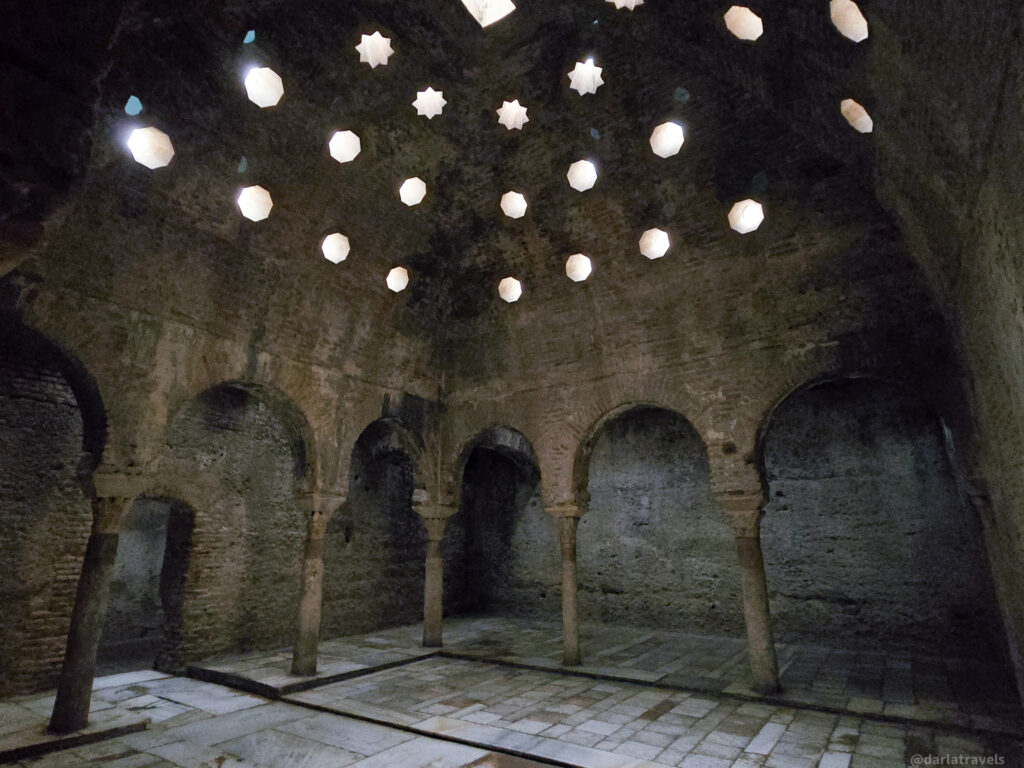 A photo of the interior of a historic bathhouse. The room has a vaulted ceiling with small, round openings that allow light to filter in. There are arches and columns along the walls.