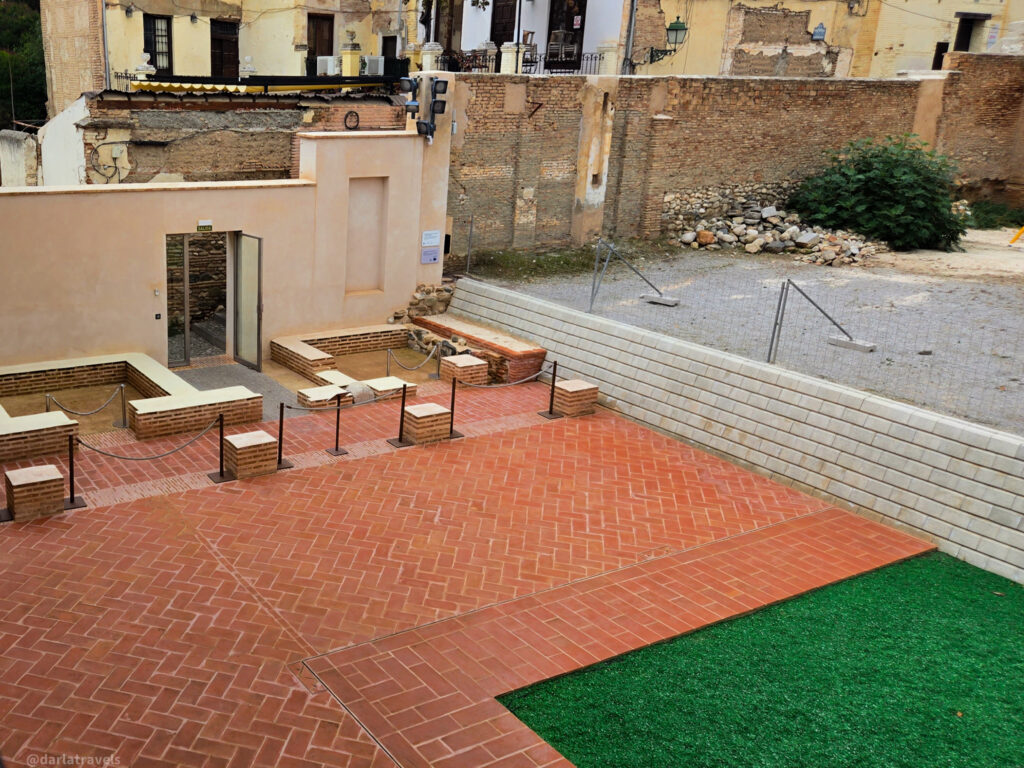 A photo of a courtyard with exposed brick walls and a partially excavated area with ancient stonework. There is a brick walkway and a patch of green grass in the foreground.