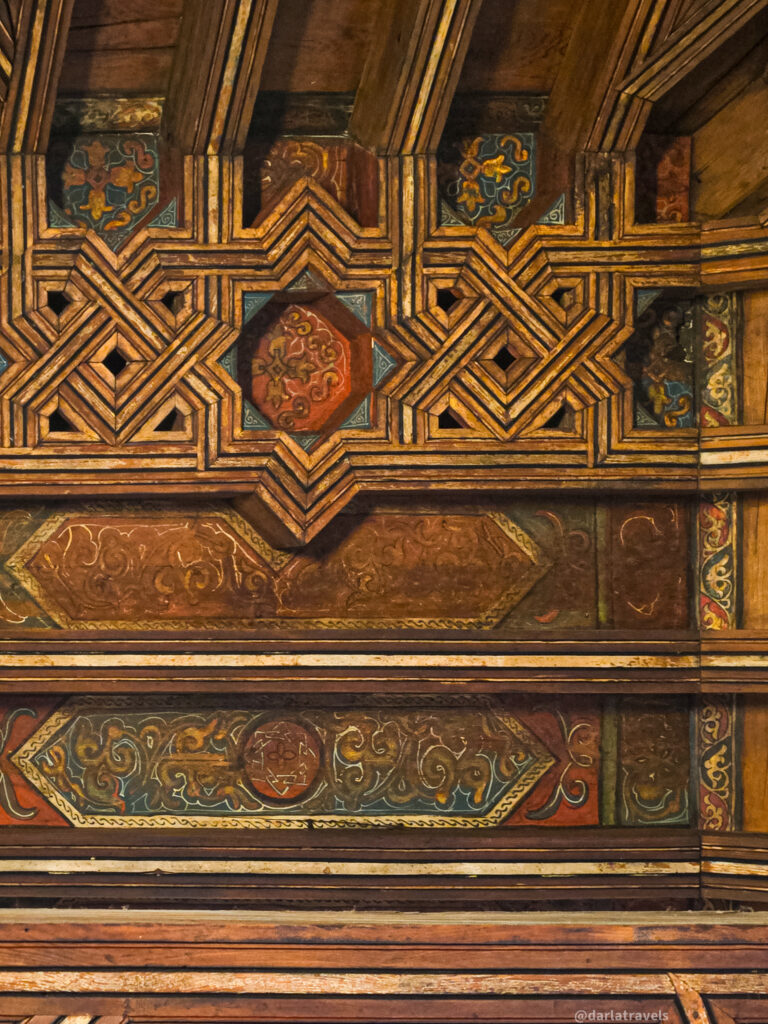 A close-up of an ornately decorated ceiling. The wooden beams are painted with intricate geometric and floral patterns in shades of brown, red, and gold.