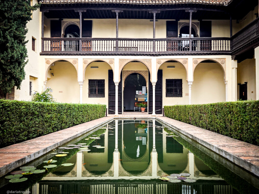 A photo of a courtyard with a rectangular pool in the center. The pool is surrounded by a two-story building with a covered walkway supported by stone columns. There is a hedge of green bushes on either side of the pool.