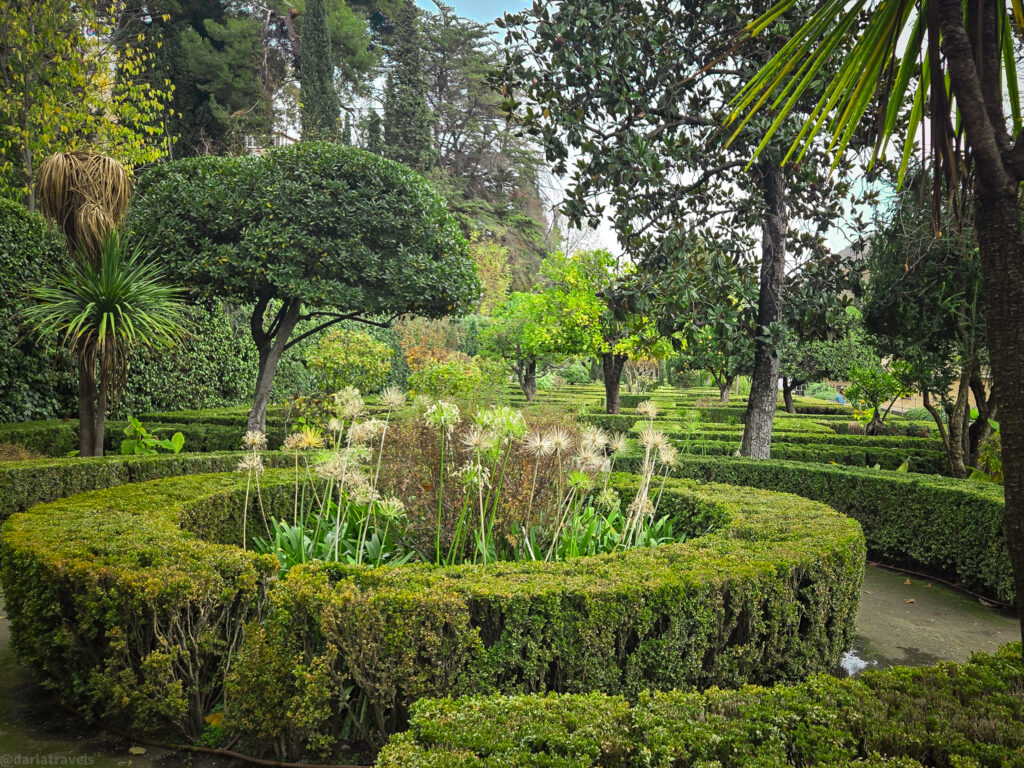 A photo of a beautifully landscaped garden with a variety of trees, shrubs, and flowers. The garden features intricate hedges forming curved paths and circular beds.
