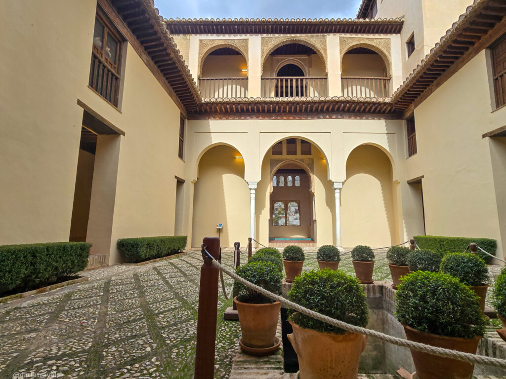 A photo of a courtyard with a two-story building surrounding it. The lower level has a covered walkway with arched doorways and columns. The upper level has a balcony with a wooden railing.