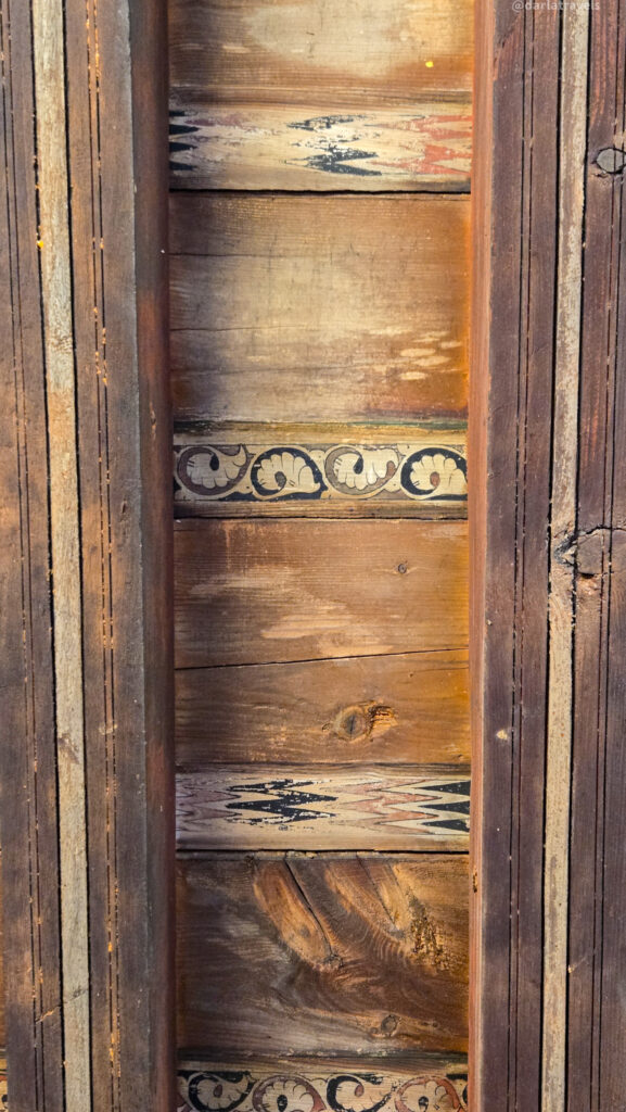 A vertical image of a wooden wall with horizontal planks. The wood has a rich, aged patina, and there are painted floral designs in blue, red, and yellow along the planks.