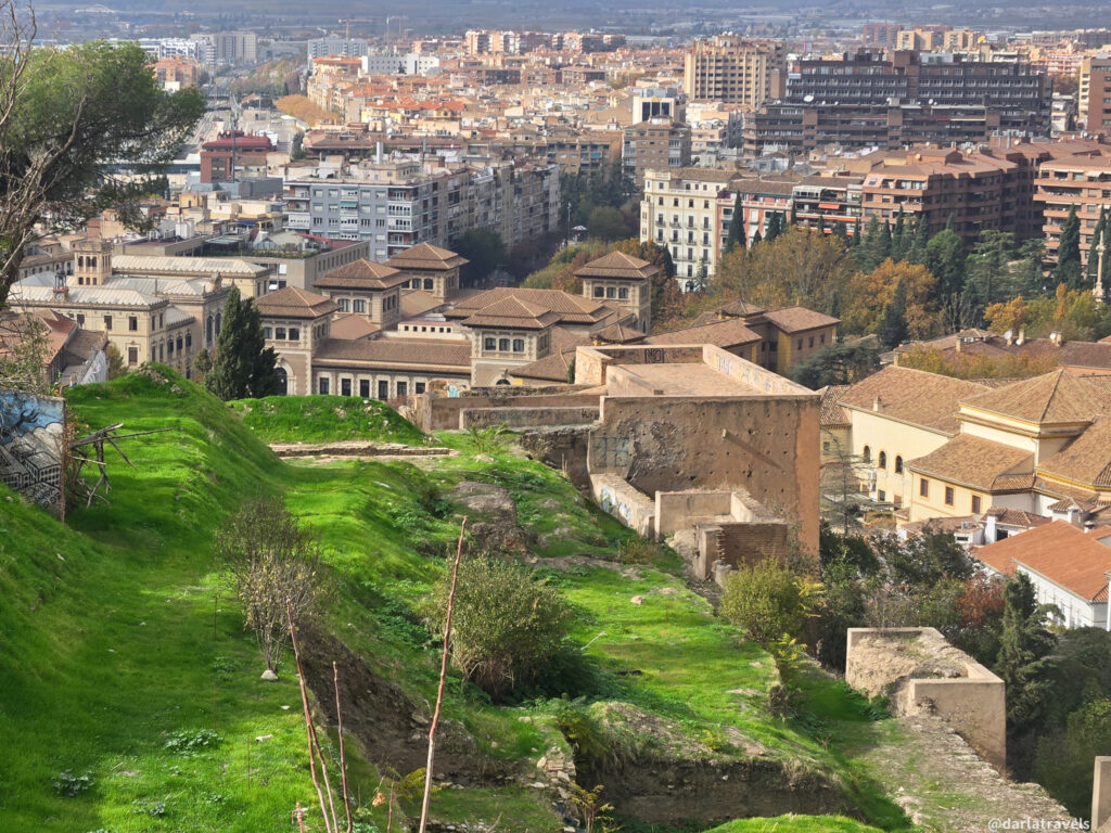 A photo showing a sweeping view of Granada, Spain. The image highlights the ancient walls and structures, nestled amidst lush greenery and overlooking the city's urban landscape.