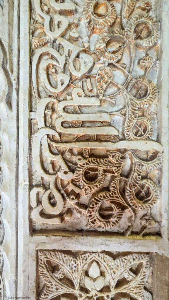 A close-up of a wall with intricate carvings and patterns. The design features geometric shapes and flowing lines, Arabic script.
