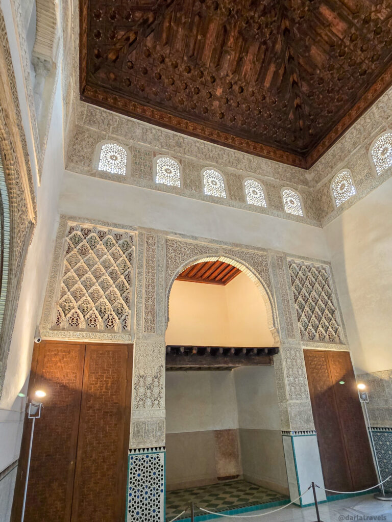 A photo of a room with a coffered wooden ceiling and intricately carved arches. The walls are decorated with geometric tilework and there are wooden doors and windows.