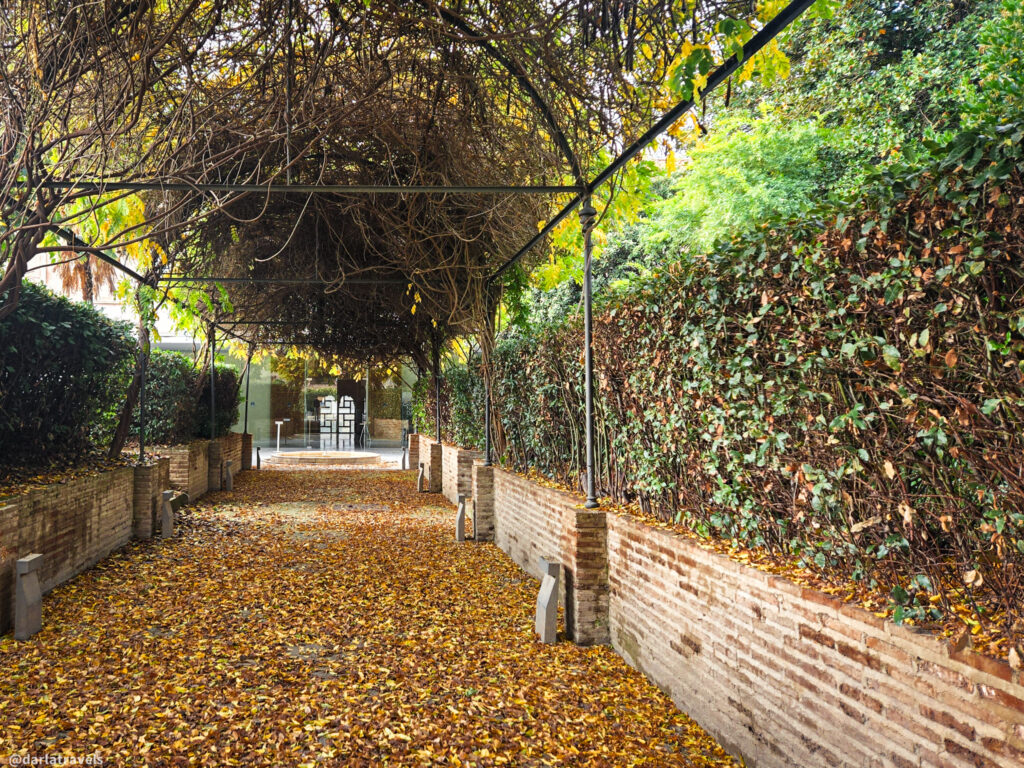 A photo of a path lined with brick walls and covered by a canopy of yellow leaves. The path leads to a doorway in the distance.