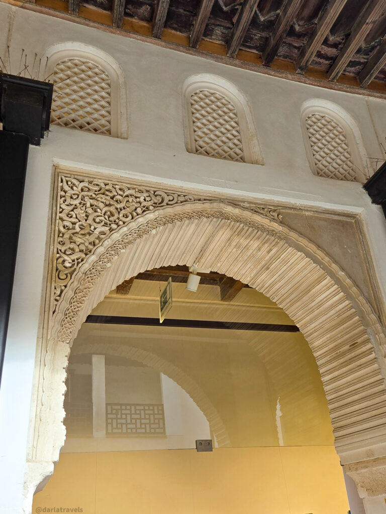 A close-up of an arched doorway with elaborate plasterwork details. Above the arch are three small windows with latticework.