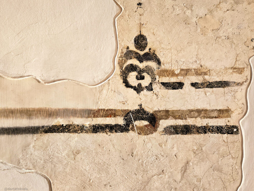 A close-up of a wall with an abstract geometric pattern painted on it. The design features shapes and lines in shades of brown.