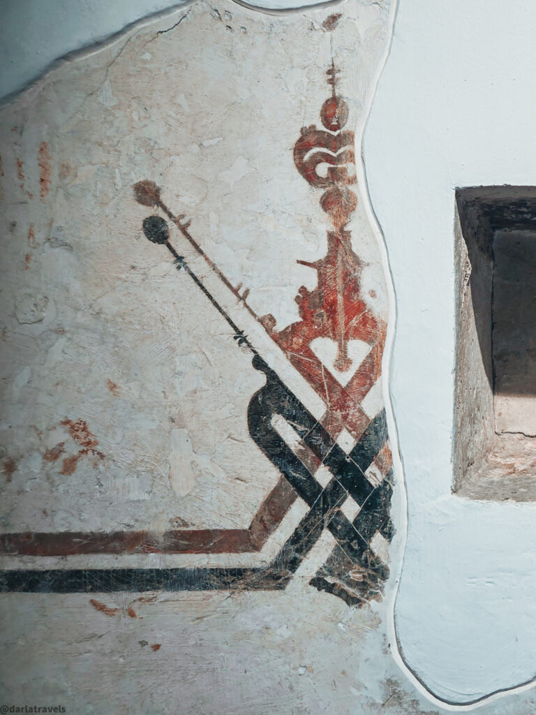 A close-up of a wall with an abstract geometric pattern painted on it. The design features shapes and lines in shades of red, brown, and black.