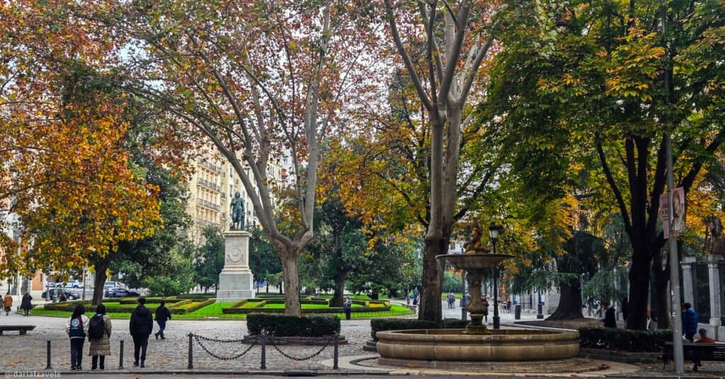 park benches looking across a street with large trees and a statue in a plaza; walks in Madrid's parks