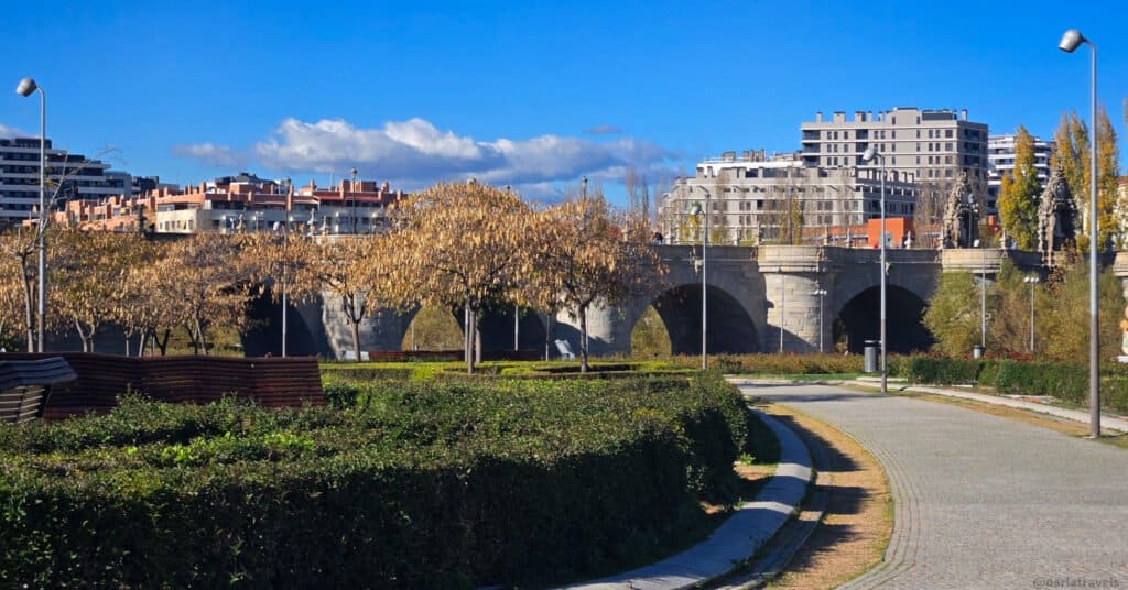 park with manucured gardens. trees obscure an ancient stone bridge with several arches