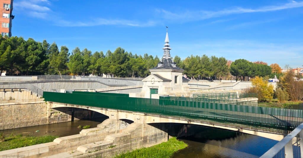 a pedestrian bridge over a river with a small decorative dam control building in the center