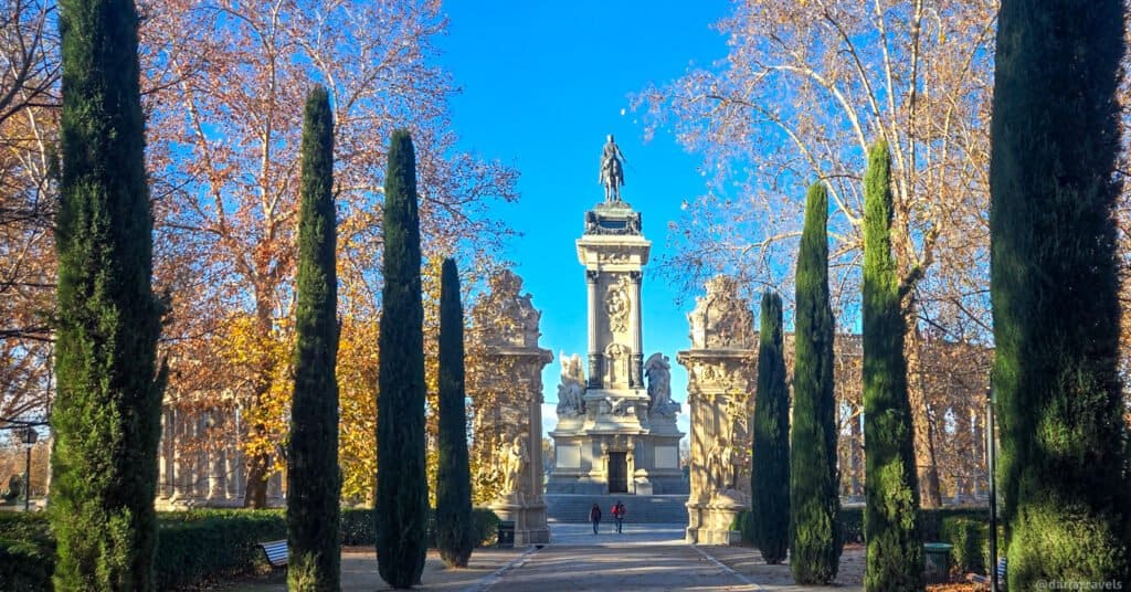 a gravel path lined with tall, narrow cypress tress leads to a large monument with an equestrian statue on top in Retiro Park Madrid