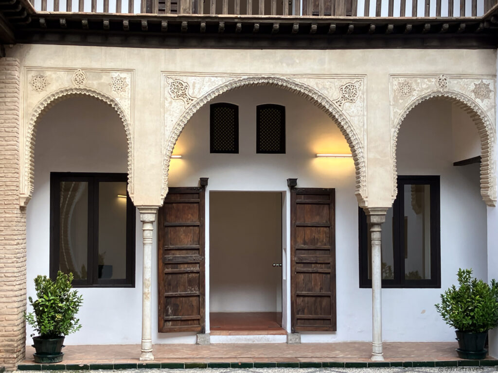 A photo of a courtyard with a two-story building surrounding it. The lower level has a covered walkway with arched doorways and columns. The upper level has a balcony with a wooden railing.
