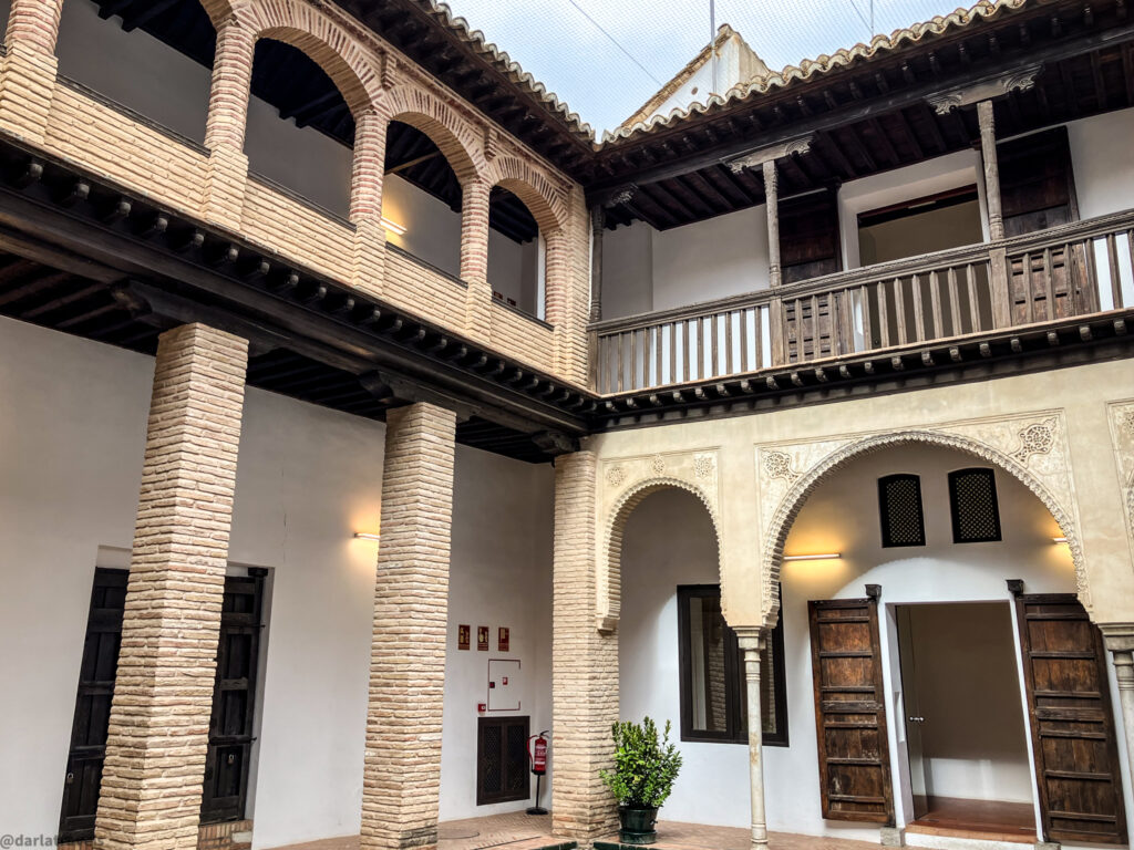 A photo of a courtyard with a two-story building surrounding it. The lower level has a covered walkway with arched doorways and columns. The upper level has a balcony with a wooden railing.