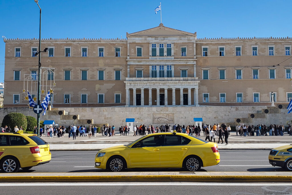 A sunny day view of the Hellenic Parliament building, a large neoclassical structure with a central colonnade. A crowd of people walks in the square in front, and several yellow taxis are in the foreground. The Greek flag is prominently displayed.