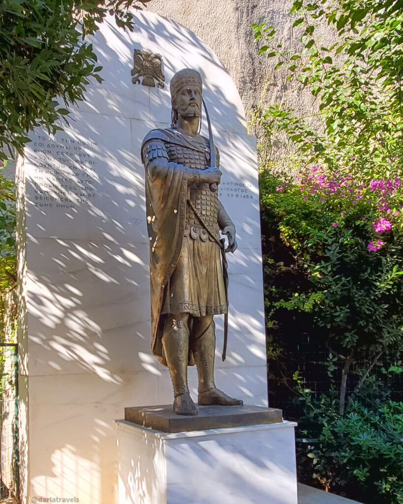 Bronze statue of Constantine I, dressed in Roman military attire, holding a staff, set against a white wall with inscriptions and surrounded by trees and flowering bushes.  On a DIY shore excursion of Athens, Greece