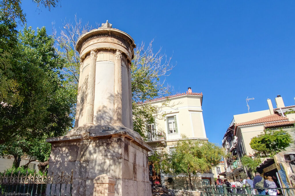 Choragic Monument of Lysicrates, a small, circular structure with a dome-like roof, located in a square in Athens. The monument is framed by trees and buildings, including a multi-story building with balconies and a red-tiled roof. People are visible in the background.