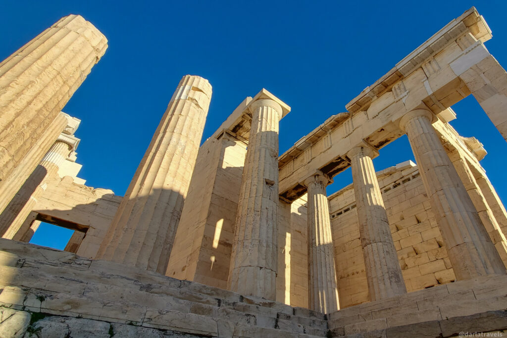 A low-angle perspective of the Propylaea, or entrance hall at the top of the Acropolis, showcasing its massive Doric columns and weathered stonework against a clear, deep blue sky. The ruins show signs of age and wear. 