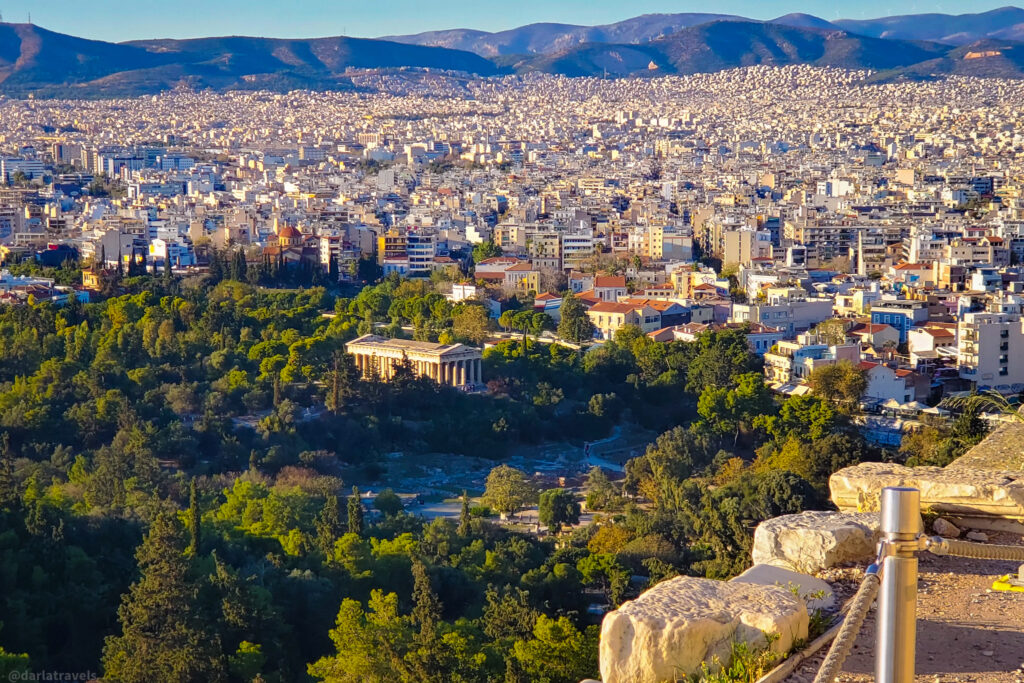 panoramic view of Athens, Greece, from an elevated vantage point. The cityscape is densely packed with buildings stretching towards the mountains in the background. In the foreground, lush greenery surrounds the well-preserved Temple of Hephaestus, an ancient Greek temple with classical columns. The warm sunlight enhances the contrast between the urban landscape and the natural elements, creating a stunning visual of the city's historical and modern blend.  Self-guided tour