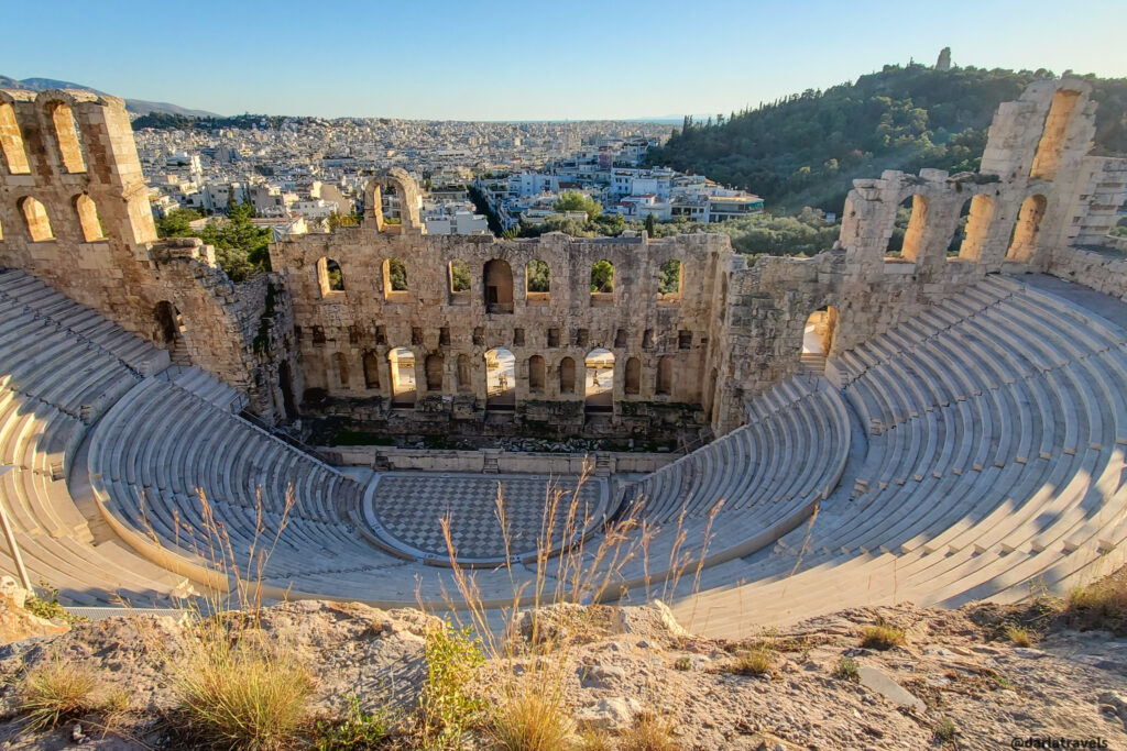 A wide shot of the Theatre of Dionysus, an ancient amphitheater in Athens, Greece, considered the birthplace of Greek tragedy. The stone seating and stage ruins are clearly visible. The cityscape of modern Athens stretches out in the background, with a hill and trees visible to the right. The late afternoon sun casts a warm light on the scene. Dry grass and foliage are visible in the foreground.