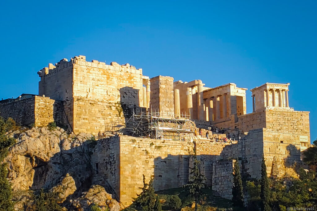 A low-angle shot of the Acropolis of Athens, showcasing the ancient ruins on a rocky hilltop against a clear blue sky. The temple of Athena Nike is prominent. A portion of the Erechtheion with its Porch of the Caryatids is visible to the right. Green trees line the base of the hill.  On your own tour in Athens, Greece