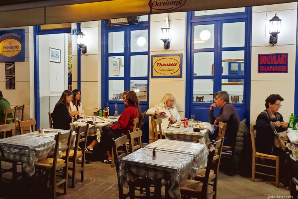 "The image shows an outdoor seating area of a restaurant named 'Thanasis Souvlaki.' The restaurant has blue-framed windows and doors, with signs displaying the name 'Thanasis' in Greek and English. Several tables are set up with checkered tablecloths, and people are seated at the tables, eating and conversing. The scene is well-lit, suggesting it is evening. The restaurant appears to be a casual dining establishment, and the setting is cozy and inviting