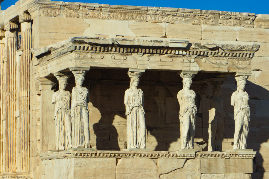 A close-up shot of the Porch of the Caryatids on the Erechtheion, located on the Acropolis in Athens. The six caryatid statues, sculpted female figures draped in cloth, stand in a row supporting the porch roof. The stone shows signs of age and weathering. See Athens on your own.