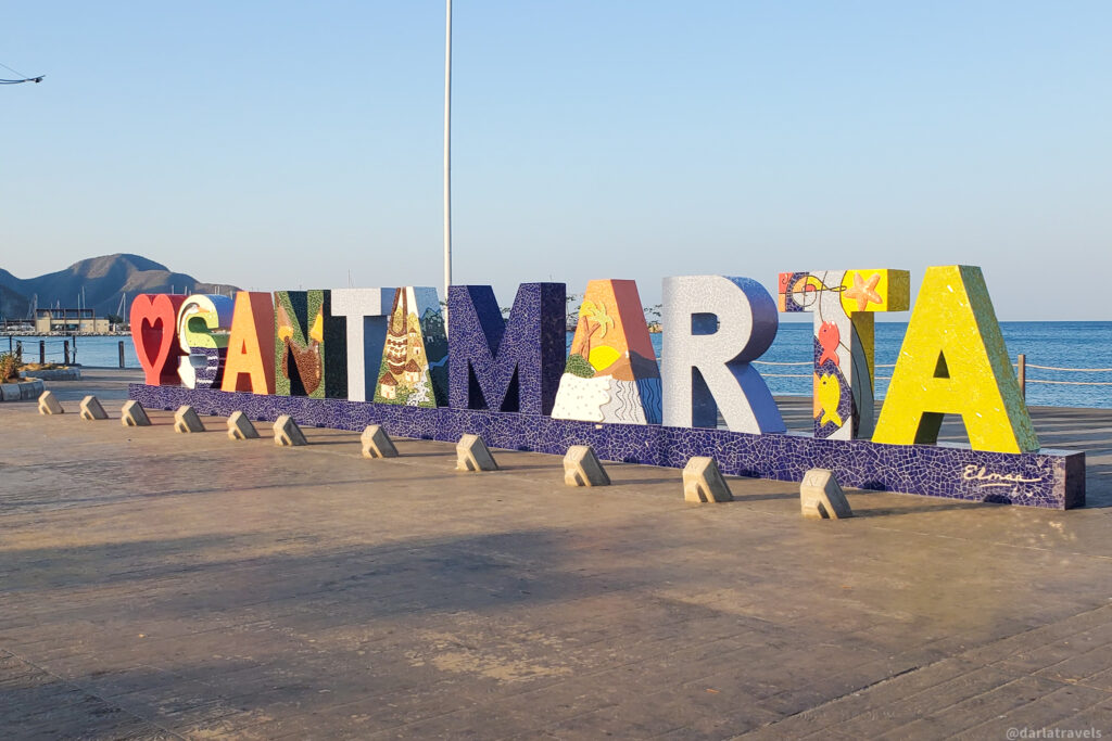 A colorful "Santa Marta" sign stands on a paved waterfront promenade, with the ocean and mountains in the background. Each letter is decorated with vibrant mosaic artwork representing local culture, nature, and marine life. The "S" is painted in green, yellow, and red, while the "A" features a church design. The "R" has a textured gray pattern, and the final "T" is bright yellow with fish and starfish illustrations. The blue sky and calm sea add to the scenic coastal atmosphere.