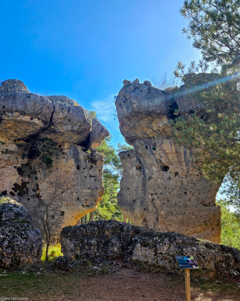 A medium shot of Los Amantes de Teruel (Teruel's Lovers), two distinct, tall rock formations in the Enchanted City near Cuenca, Spain. The rocks are shaped in a way that resembles two faces looking towards each other. The background features trees and a bright blue sky. The foreground includes rocks and dry ground, with a small sign on a wooden post visible.